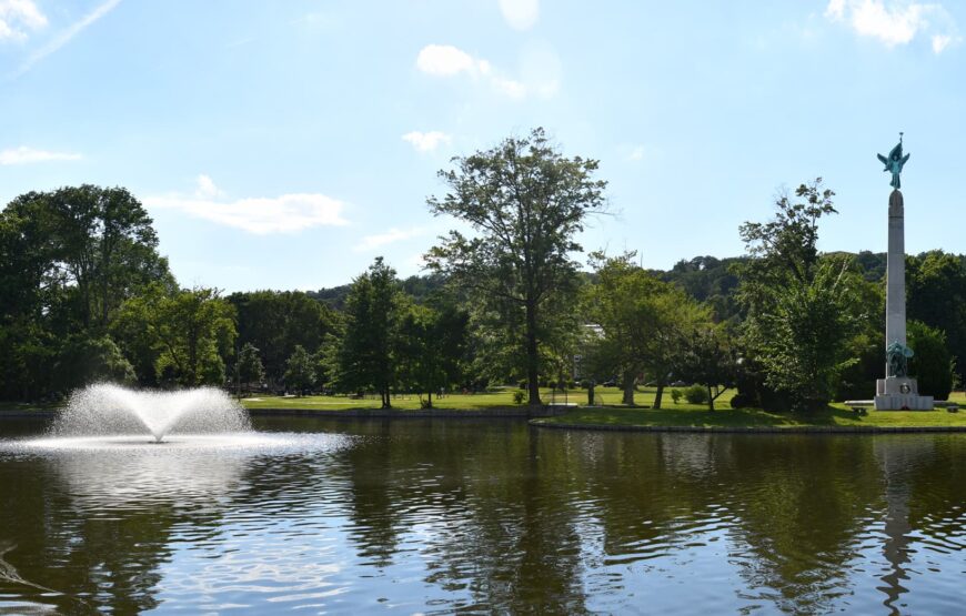 View of Edgemont Memorial Park lake in Montclair, New Jersey, featuring a water fountain, tree-lined shoreline and the World War I memorial monument on a sunny day.