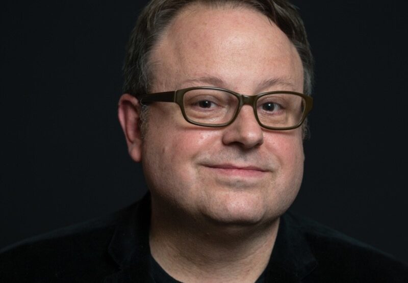 Tom Hall, artistic director of Montclair Film, smiling in glasses and a dark blazer against a black background